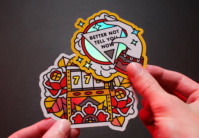 2 luck-themed hologrpahic stickers with 'Better Not Tell You Now' text held by a hand against a dark background