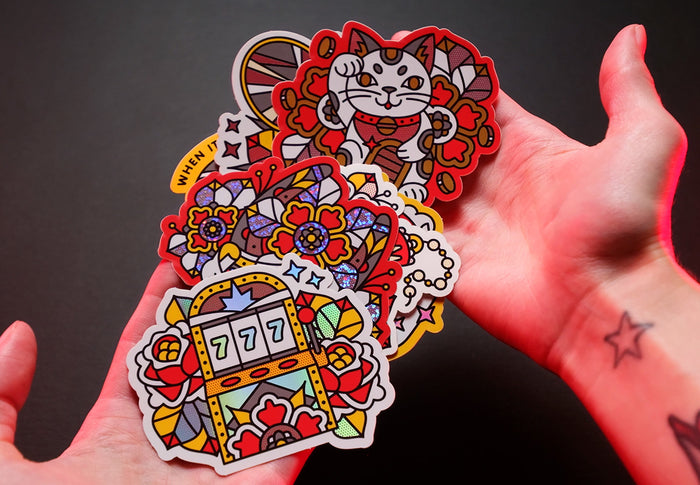 Colorful luck-themed die-cut stickers featuring a cat, slot machine, and flowers held in hands against a dark background.
