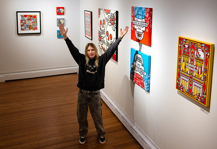 Lisa Champ of Red Halftone standing in Pratt Munson gallery with arms raised, surrounded by her colorful artworks on a white wall.