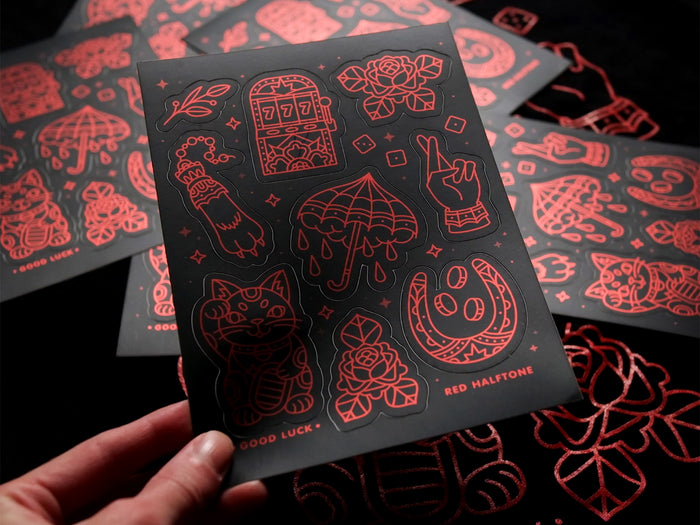 A black sticker sheet with luck-themed red line art designs held by a hand against a dark background