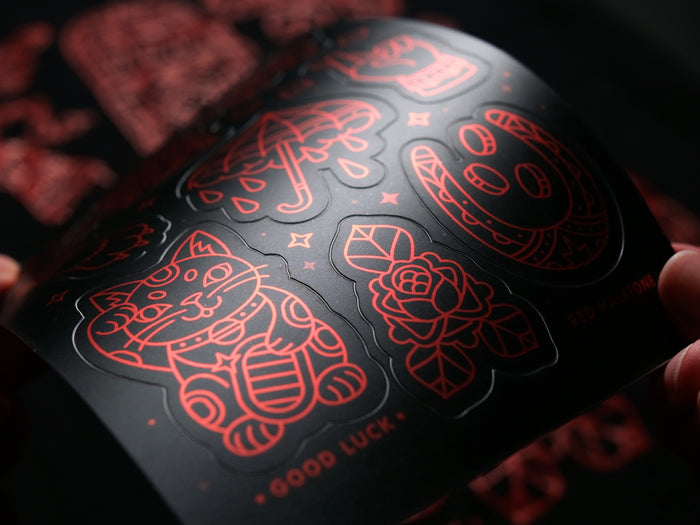 A close-up of a black sticker sheet with luck-themed red line art designs 