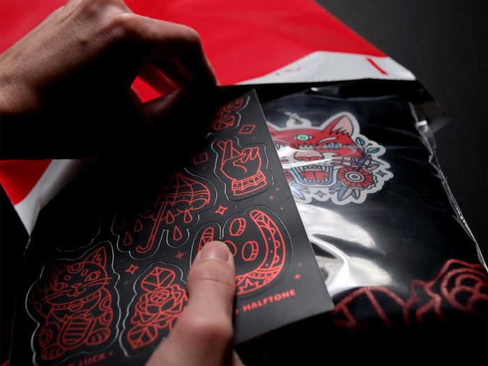 Person packing a stickersheet and black t-shirt in a red polybag against a dark background
