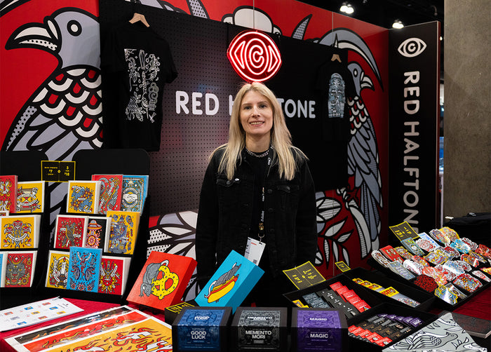 Lisa Champ of Red Halftone is standing behind her booth at Adobe MAX with various products on display.