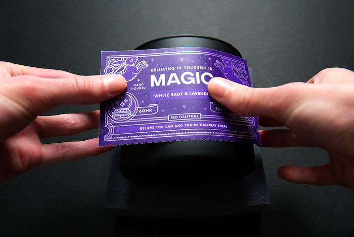 Hand applying a purple product label to a black glass jar against a dark background