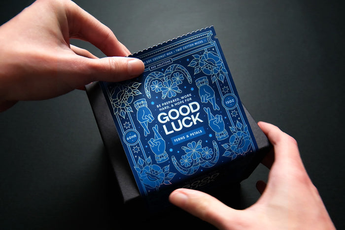Applying an ornate blue label with 'Good Luck' text to a box held by hands against a dark background