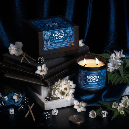 A dramatically lit scene with old books, white marble, white flowers, ferns, and dice featuring a scented candle with an illustrated blue label that reads Good Luck.