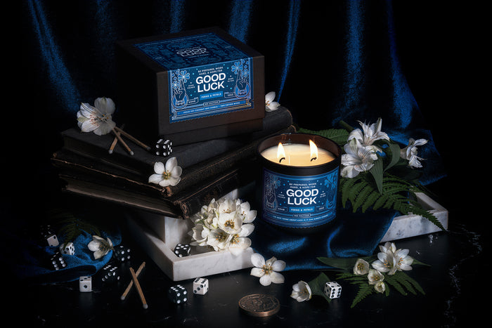 Good Luck candle and packaging by Red Halftone on blue velvet background with white flowers, ferns and dice.