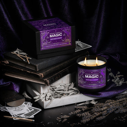 A dramatically lit scene with old books, white marble, playing cards, dried sage and lavender featuring a scented candle with an illustrated purple label that reads Magic.
