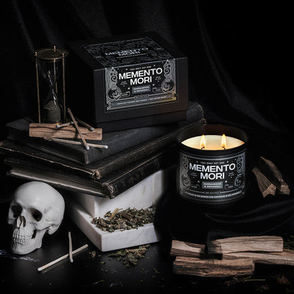 A dramatically lit scene with old books, white marble, a skull, an hourglass, and Palo Santo sticks featuring a scented candle with an illustrated black label that reads Memento Mori.