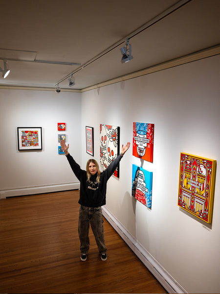 Lisa Champ at Pratt Munson gallery with arms outstretched, surrounded by her colorful artworks on white walls.