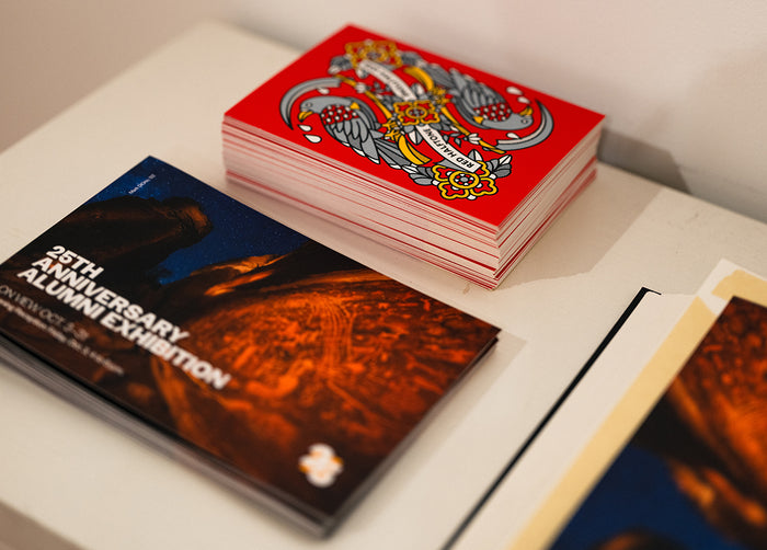 Stack of red Red Halftone postcards, and a black book with '25th Anniversary Alumni Exhibition' text below.