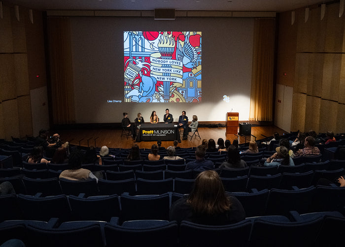 Audience in a theater watching an artist panel with a colorful slide on the screen.