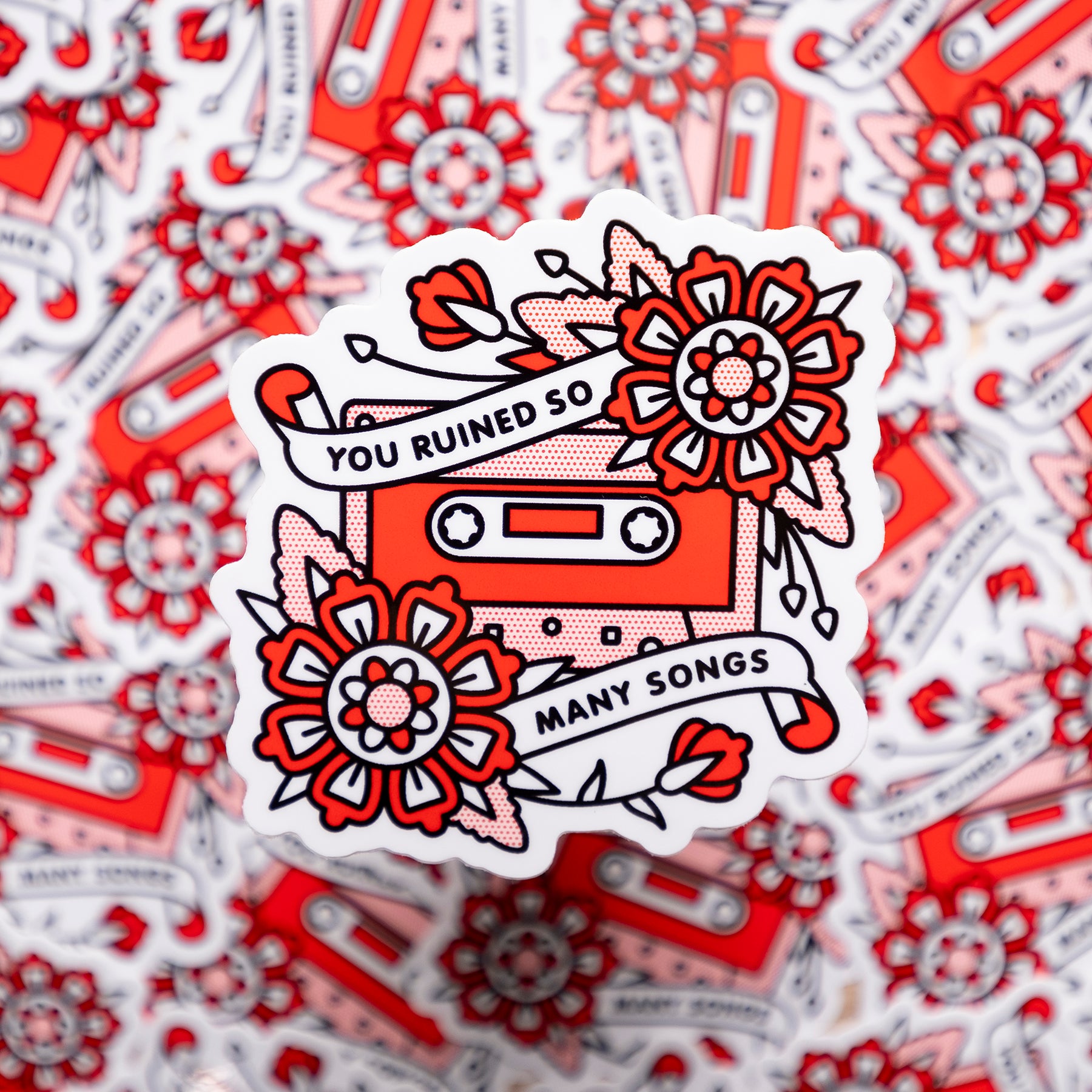 A 3.1x3.1" vinyl art sticker of a cassette tape with two flowers and banners overlaid on adjacent corners with text that reads: You ruined so many songs. Drawn in a flash tattoo and pop art fusion style in a red, white and black color palette by the artist, Red Halftone.